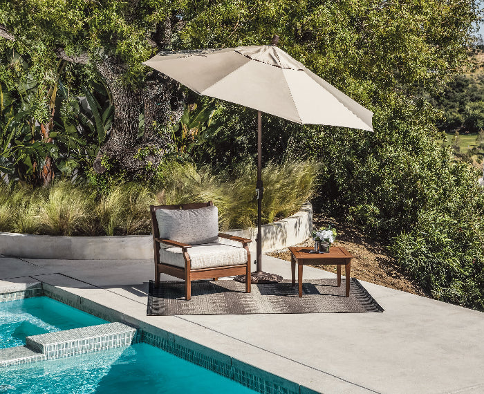 Outdoor seating area with a chair, table, and umbrella by a pool surrounded by greenery.